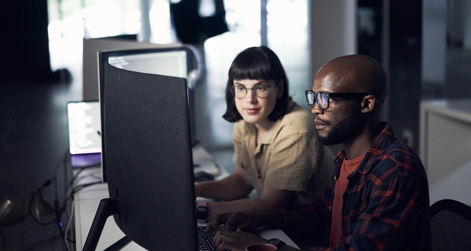 Developers, coding and programming at night on computer in software or application development at office. Man and woman code on desktop PC working late in team testing, security or online networking Kommunikatörer framför dataskärm
